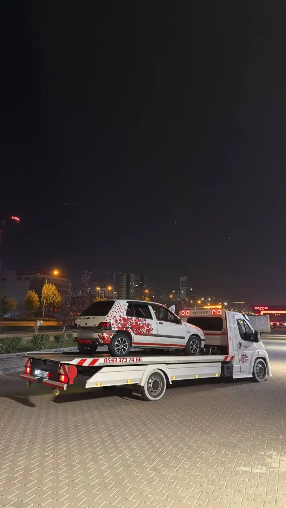 Birlik Oto Kurtarma çekicisi gece, şehir ışıkları önünde kasasında beyaz bir hatchback taşıyor